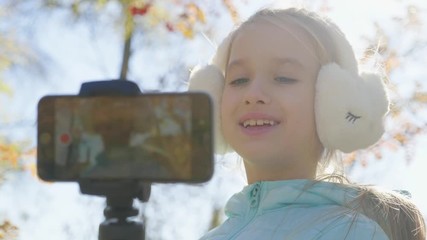 Happy cute little vlogger making online video recording vlog sitting on a park bench. Record vlog and blog concept for Internet.