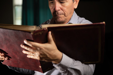 Mature man with glasses at home watching an old photo album remembering the past and looking at the...