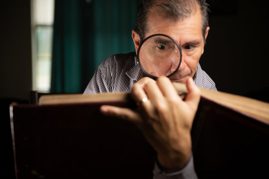 Mature man with glasses at home watching an old photo album remembering the past and looking at the pictures with a magnifying glass