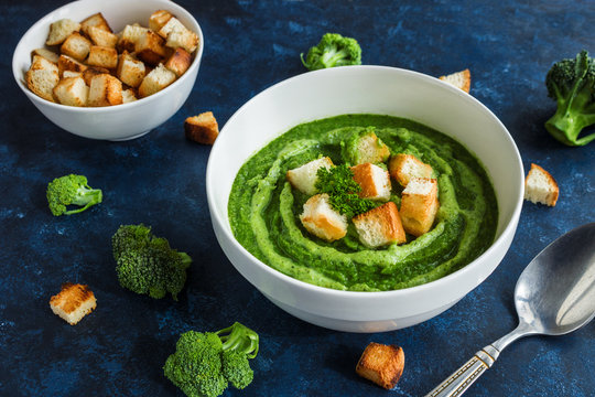 healthy broccoli soup with croutons in bowl tablespoon and raw ingredients on dark blue 