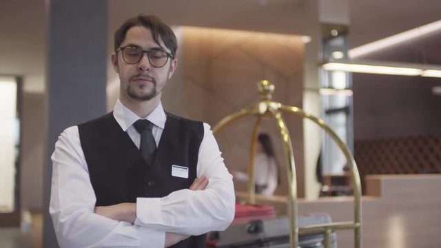 Tracking portrait shot of professional bellman or concierge standing in lobby of hotel with his arms crossed and looking at camera