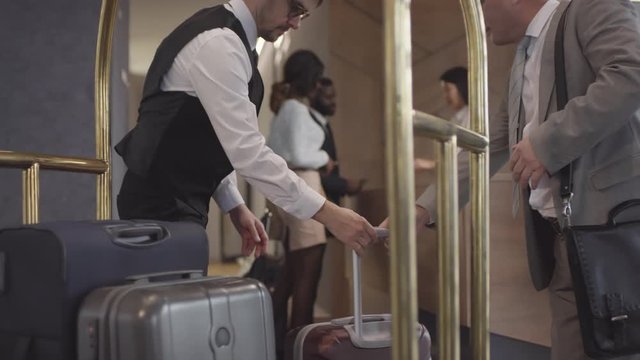 Tracking shot of professional hotel porter in glasses and uniform helping guests and putting their suitcases on luggage cart, then pushing it and leaving