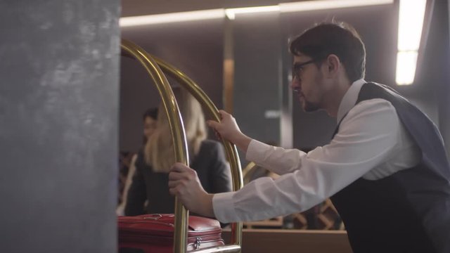 Tracking shot of serious bellhop in uniform pushing luggage cart down hotel hallway