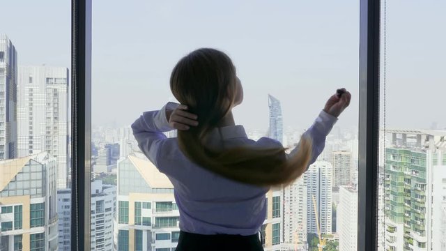 Back View of Business Woman with Long Hair Take Off the Scrunchy and Lightly Shakes Her Hair After Hard Day Near Window with Modern City with Skyscrapers Buildings on Background. Slow Motion