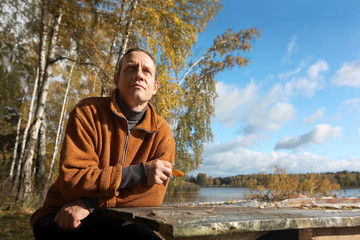 A man in an orange sweater is sitting on the street at a table from a thick board. Autumn forest