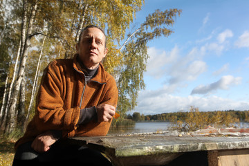 A man in an orange sweater is sitting on the street at a table from a thick board. Autumn forest