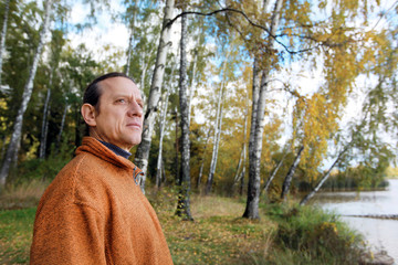 A man in an orange sweater against the background of a river bank with a birch grove. Autumn forest