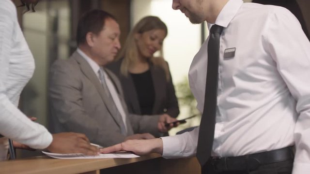 Tracking shot of male and female receptionists checking guests into hotel. They are receiving payment, handing room key cards and giving registration form to fill out