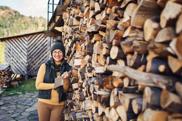 cheerful nerd girl in glasses stands with book on outdoors near stack of firewood.