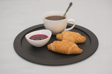 Morning coffee in a white cup with croissants and raspberry jam on a black tray.