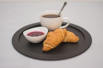 Morning coffee in a white cup with croissants and raspberry jam on a black tray.
