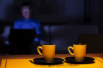 Two cups of coffee on a table and man with laptop on a couch in the background