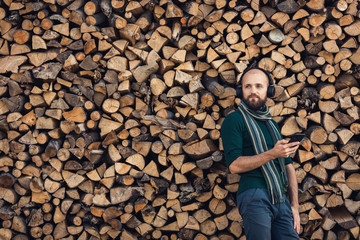 bearded man in bluetooth headphones listening to music outdoors near pile of firewood.