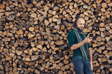 bearded man in bluetooth headphones listening to music outdoors near pile of firewood.