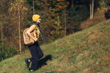 Young tourist girl with backpack rises in autumn on hill in forest in mountains.