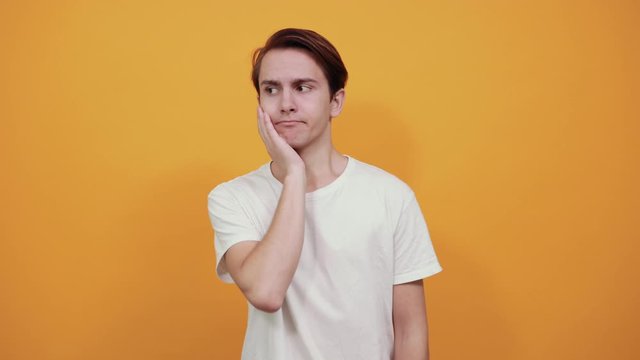 A perplexed young man in white t-shirt looks ahead in confusion, leaning head on hand against yellow background