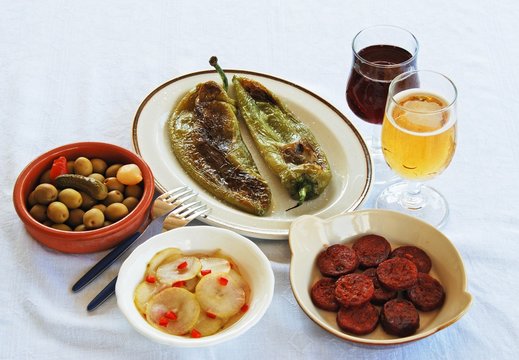 A selection of tapas with red wine and beer, Fried pointed green peppers, Green olive cocktail, Pickled cucumber with chilli, and fried Chorizo slices, Spain.