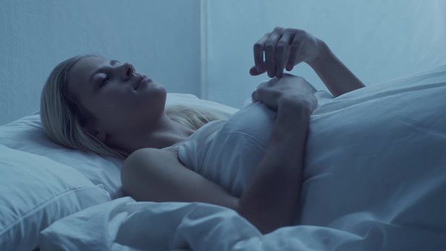 Young woman lying in the bed at night and having insomnia disease. Beautiful blond sleeping girl. Twilight in the bedroom, moonlight from the window. Sleeplessness concept.