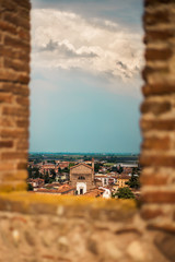Vertical view on roofs of Bergamo, Lombardy, Italy thrught embrasure from red bricks. Cloudy light blue sky above red roofs of medieval city. City landscape with rooftops and spring blooming trees