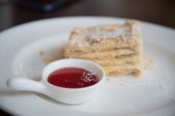A piece of Napoleon cake on a white plate. The cake is sprinkled with powdered sugar. There is a Cup of jam next to it.