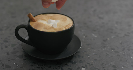 adding marshmallow into cappuccino in black cup on terrazzo countertop