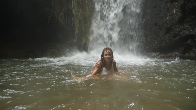 Wide shot of a young woman, dancing in the water with a yellow piece of fabric in slow motion. She is standing in a pool of water from a small waterfall behind her.