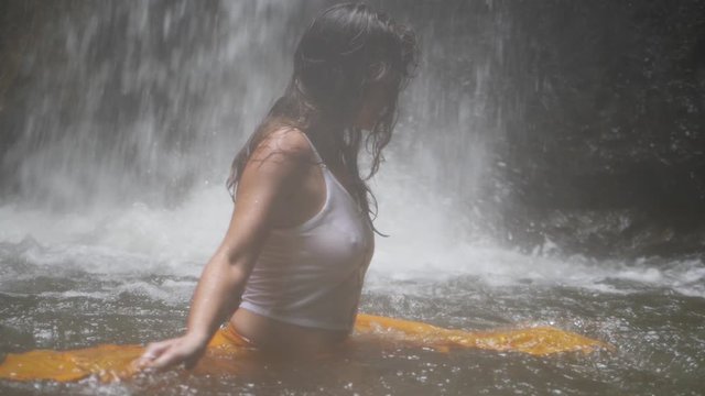 A closeup shot of a young woman, dancing in the water with a yellow piece of fabric in slow motion. She is standing in a pool of water from a small waterfall behind her.