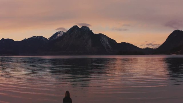 Anamorphic 4K wide aerial Drone footage of a young woman standing at the shore of the famous and scenic and idyllic Bavarian lake Walchensee in southern Germany in the alps by sunset.