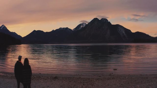 Anamorphic 4K Aerial Drone footage of a young German couple standing at the shore of the famous and scenic and idyllic Bavarian lake Walchensee in southern Germany in the alps by sunset.