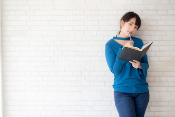 Beautiful asian woman smiling standing thinking and writing notebook on concrete cement white background at home, girl homework on book, education and lifestyle concept.