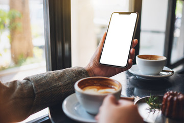 Mockup image of a woman holding black mobile phone with blank desktop screen while drinking coffee in cafe