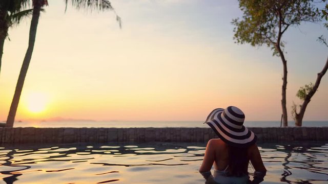 A woman with her back to the camera looks out from the comfortable resort pool waters to the colorful sunset along the ocean horizon. copy space