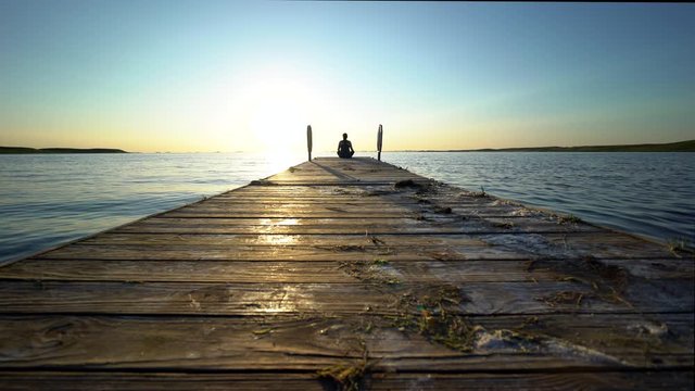 A woman sits at the end of a wooden dock while calm waves move under a beautiful sunset over Bitter Lake in South Dakota. 4k 23.98fps