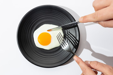 Hands are going to eat with knife and fork a fried egg on black plate on  white background.