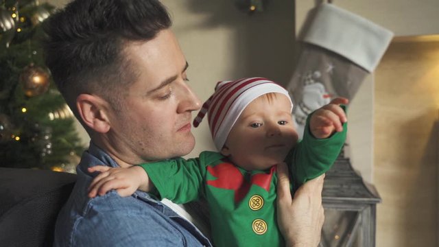 Adorable baby in elf costume wiggles in arms of daddy while sitting in front of Christmas tree. Close up, slow motion view of father holding and playing with young son.