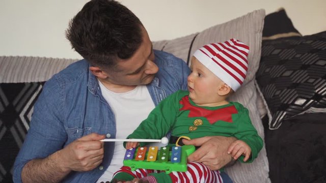 Baby in elf pajamas sitting on lap of dad looks at father's face in amazement. Happy daddy playing toy xylophone for young son on Christmas morning. Amazed adorable baby boy looks at dad.