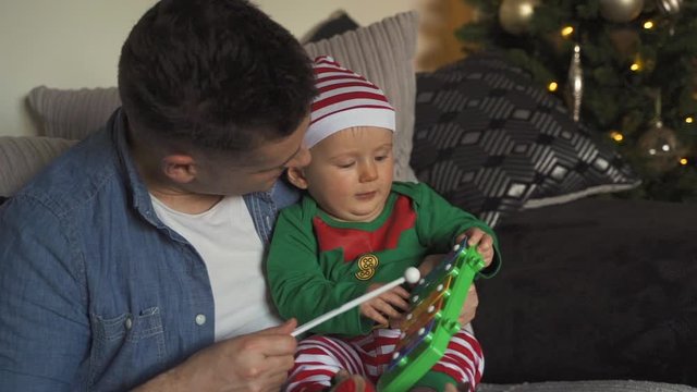 Father and child playing toy xylophone. Baby in elf costume enjoying a cuddle with daddy on the floor near the Christmas tree. Man hammering toy musical instrument. ZOOM IN.