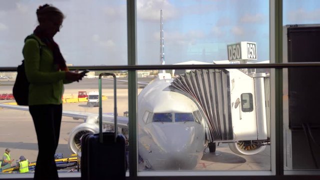 Plane is in focus as silhouette of woman at an airport terminal lounge texts on smartphone.