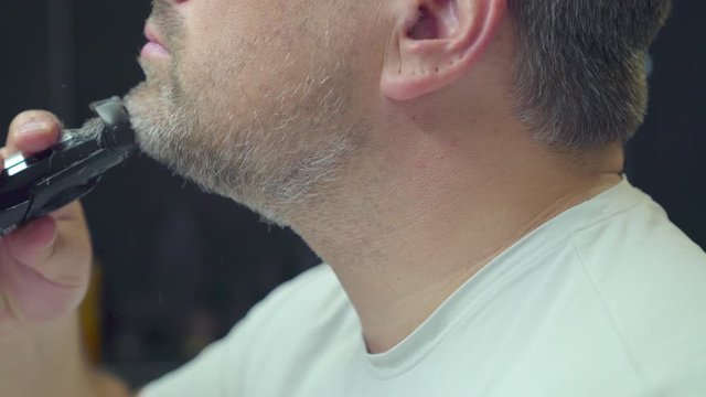 A man shaves off his beard. He has grey and black hair and shortens his beard from long to stubble