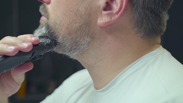 A man shaves off his beard. He has grey and black hair and shortens his beard from long to stubble