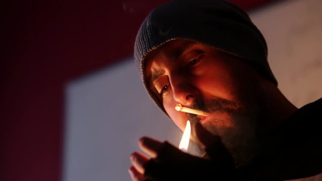 Young bearded man with hat and gloves lighting cigarette and smoking indoor in dark area with blurry background, copy space