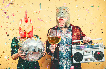 Crazy couple celebrating new year eve wearing chicken and dinosaur t-rex mask - Young trendy people having fun drinking champagne and listening music with vintage boombox - Absurd and holidays concept - obrazy, fototapety, plakaty