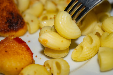 A few boiled macaroni on a fork and a white plate with fried chicken nuggets, macaroni and ketchup on a dark background. Close up.