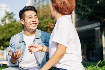 Portrait of young Asian man and woman in love sitting together on bench outdoors on sunny day chatting