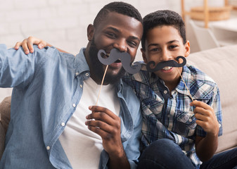 Black father and son taking selfie, holding fake moustache on sticks