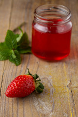 Aerial close-up of a strawberry, a recycled glass jar with strawberry jelly and mint branch, with selective focus, on rustic wooden board, in vertical