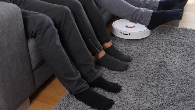 People on couch lifting their legs for a robot vacuum cleaner 