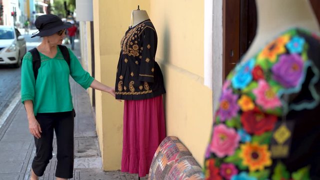 Camera focuses on Mexican blouse outside store as mature woman tourist walks up and looks at it in Merida, Mexico.