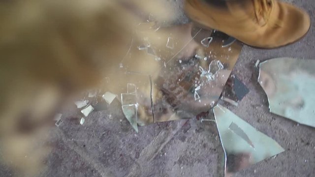 A young woman is smashing pieces of a mirror showing her own reflection with her boots on a dirty, dusty, concrete floor. Angry and furious with energy.