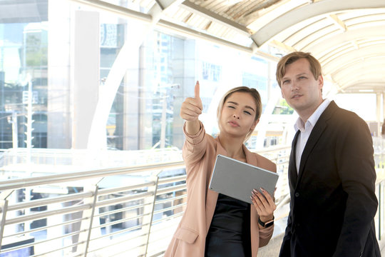 Cheerful businessman and businesswoman using digital tablet together for discussion about work while standing in the city, the lady giving thumb up to estimate size of something, businesspeople talkin